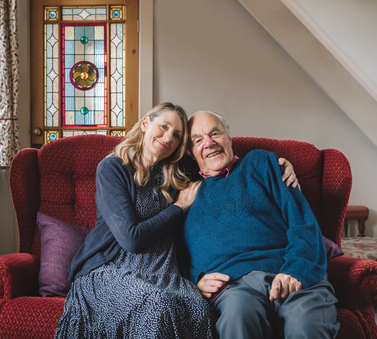 Woman with long hair sitting in a couch with her senior dad wearing blue sweatshirt and both smiling together