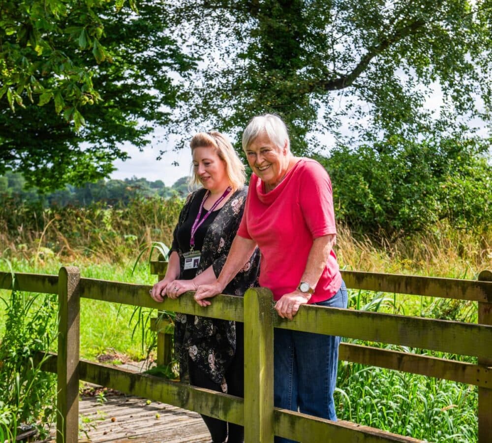 Two women standing on a wooden bridge, smiling, with green trees and grass in the background. - Home Instead