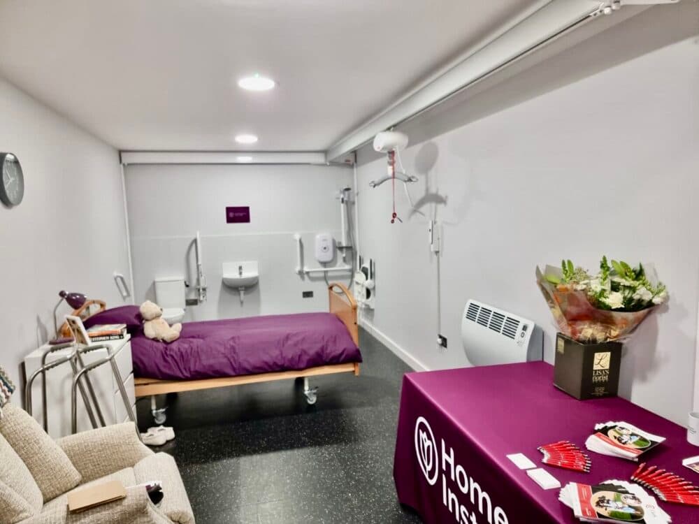 A hospital-style room with a bed, assistive equipment, a sofa, and a table holding brochures and flowers. - Home Instead