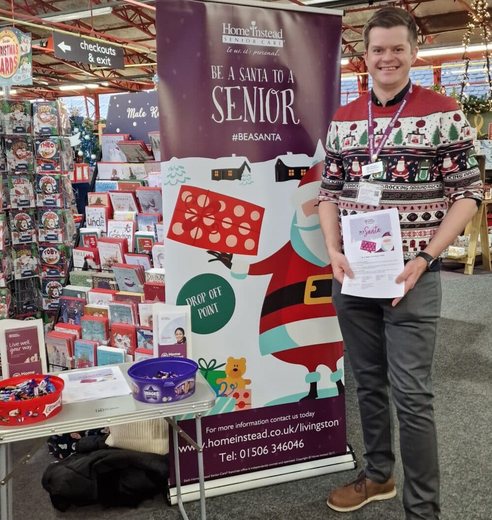 Person wearing Christmas sweater holding flyers in front of pull-up banner and table - Home Instead