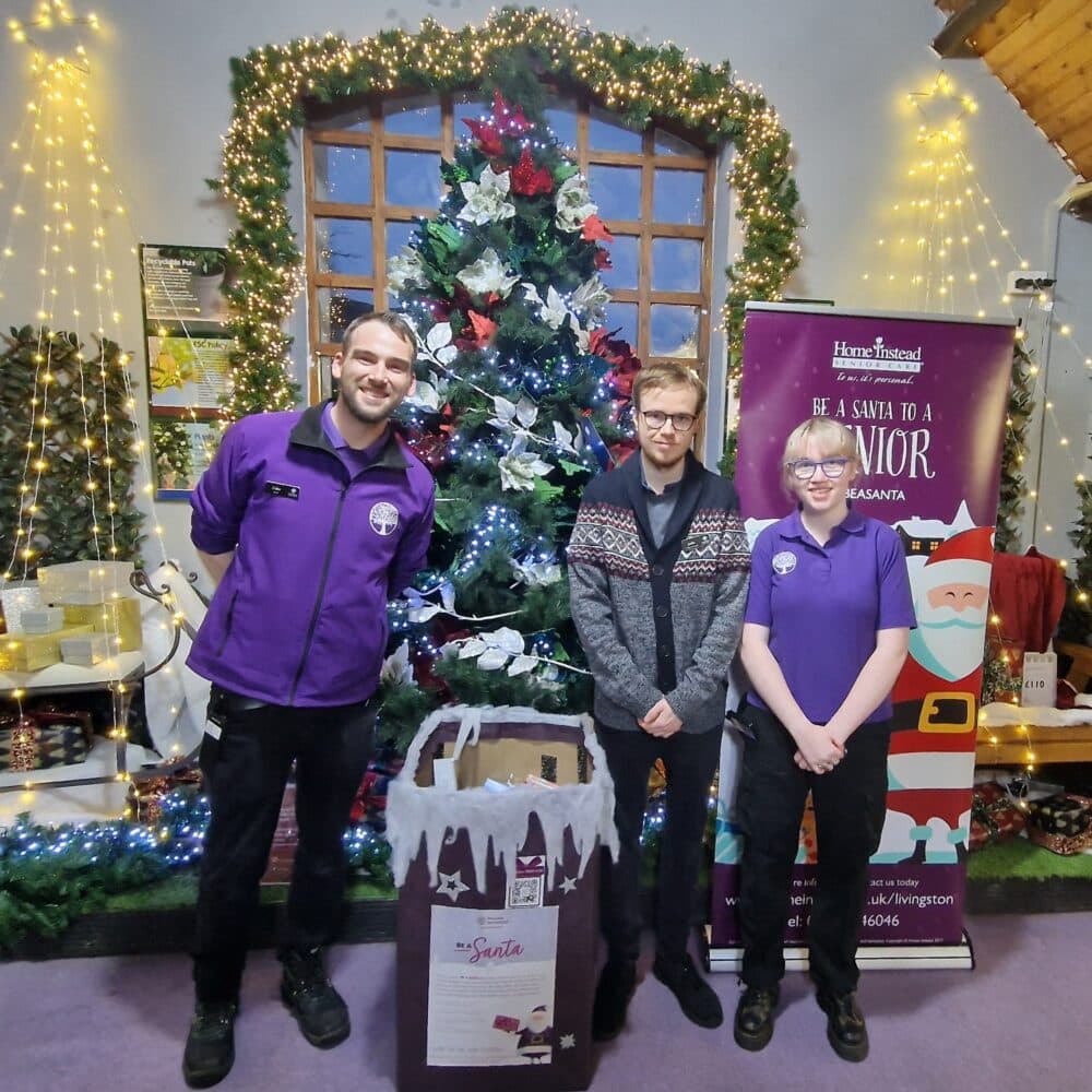 Three people in uniform standing in front of a Christmas tree with a collection bin and pull-up banner - Home Instead
