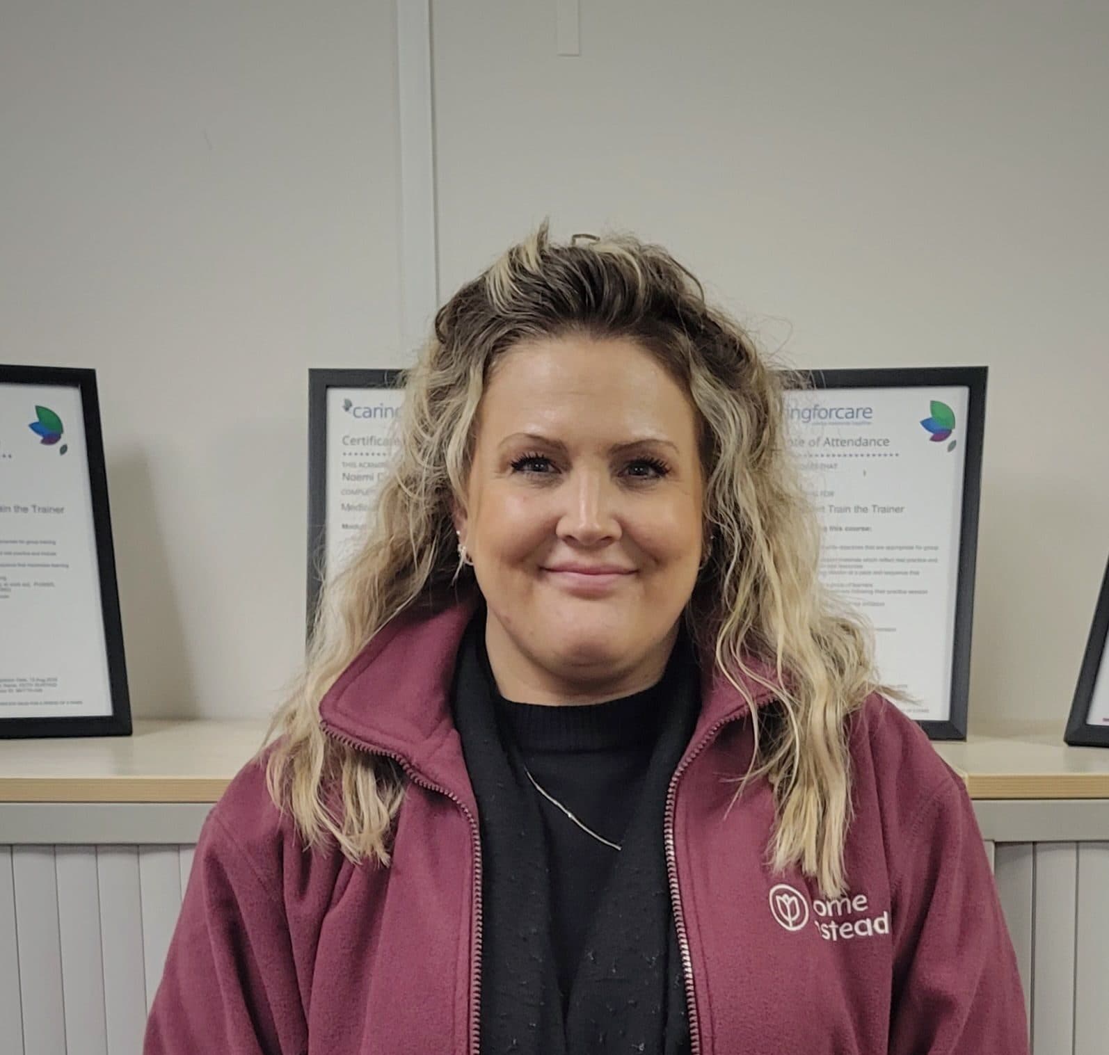 Woman with wavy blonde hair in a maroon jacket standing indoors, certificates displayed on the shelf behind her. - Home Instead
