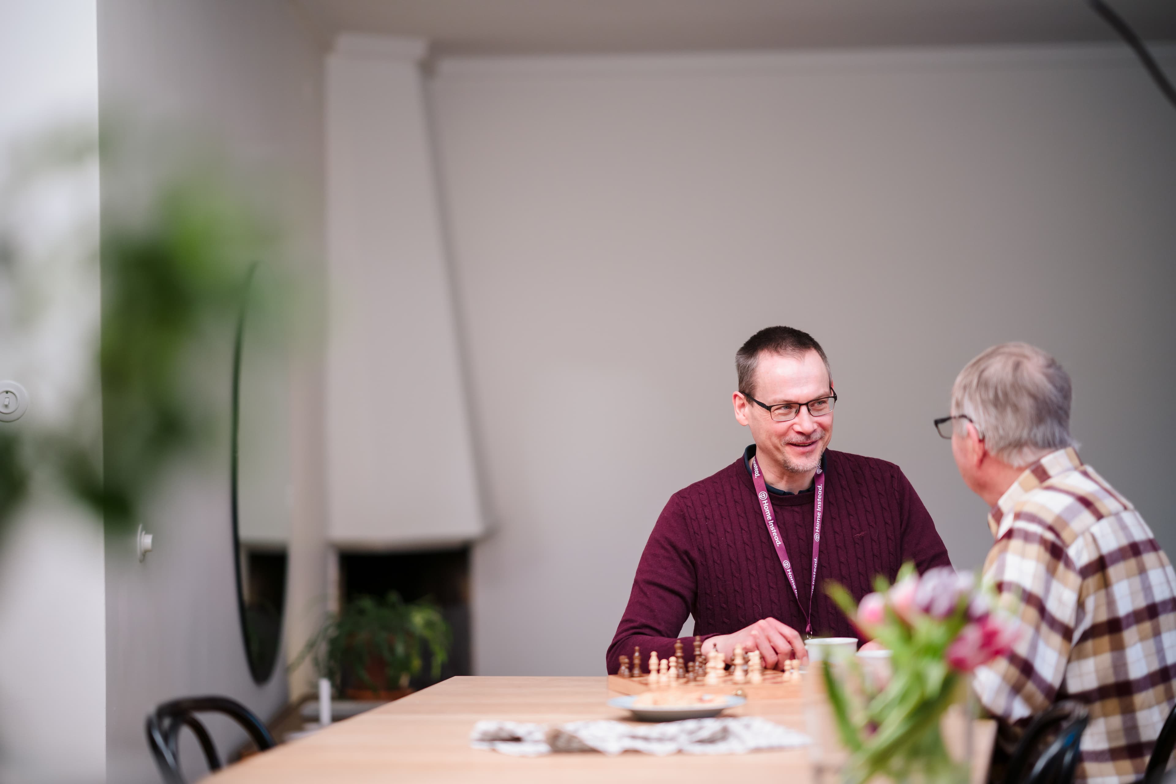 Two men sit at a table, smiling and talking, with a chessboard and flowers in the foreground. - Home Instead