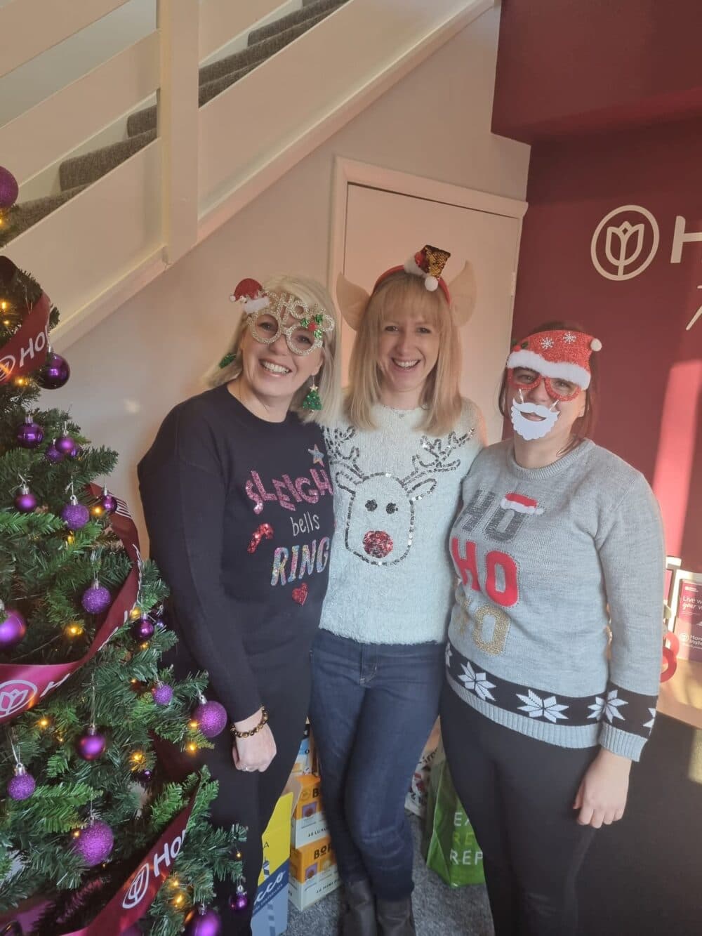 Three women in Christmas sweaters and festive accessories smiling by a decorated Christmas tree indoors. - Home Instead