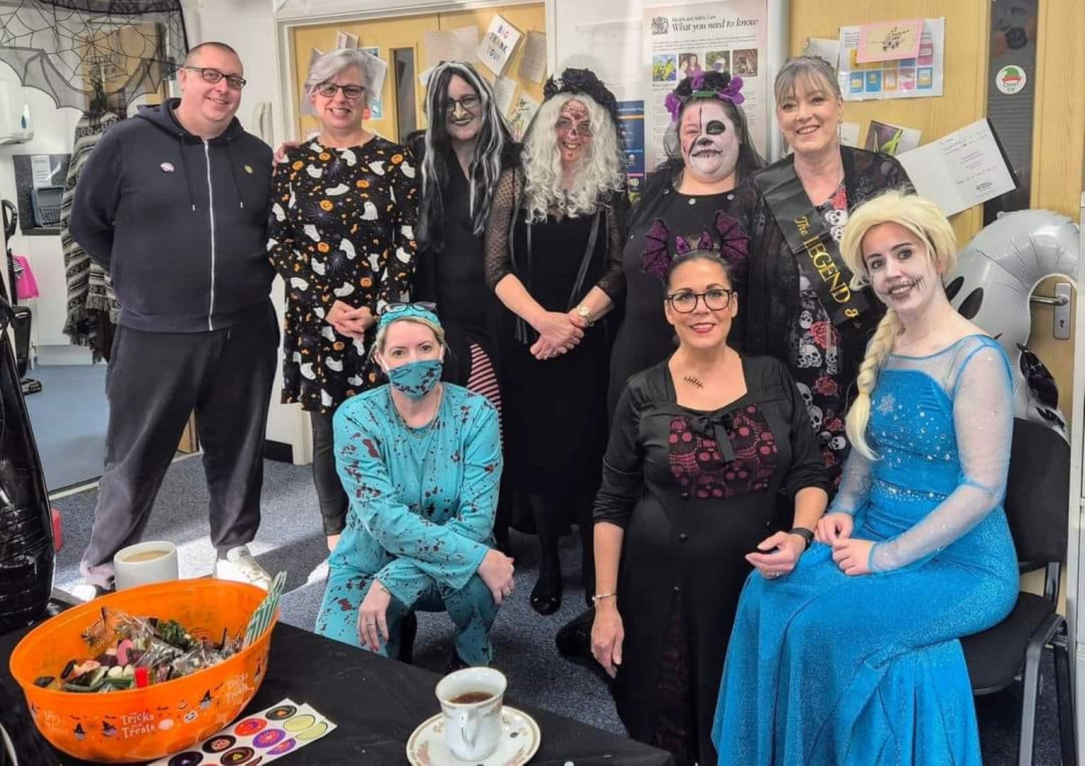 Nine adults in Halloween costumes pose indoors, with treats and drinks on a nearby table. - Home Instead