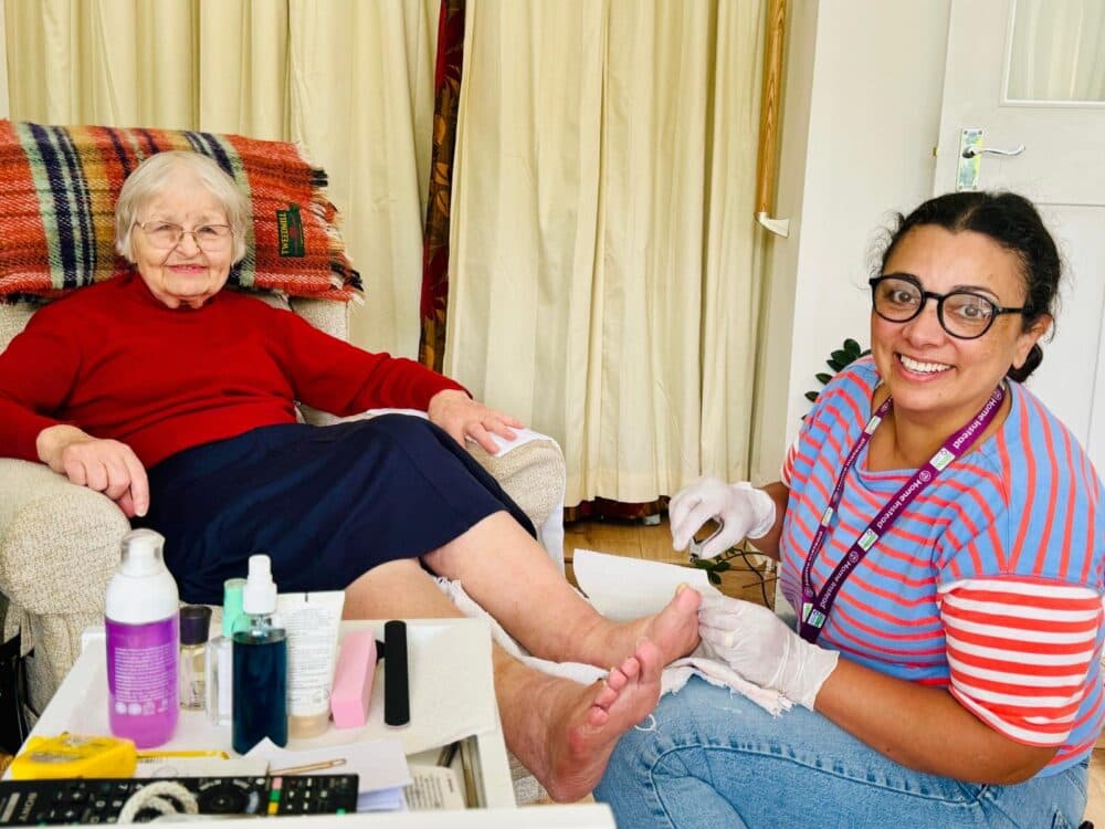 Elderly woman smiling as a caregiver gives her a pedicure at home; various supplies are on a nearby table. - Home Instead