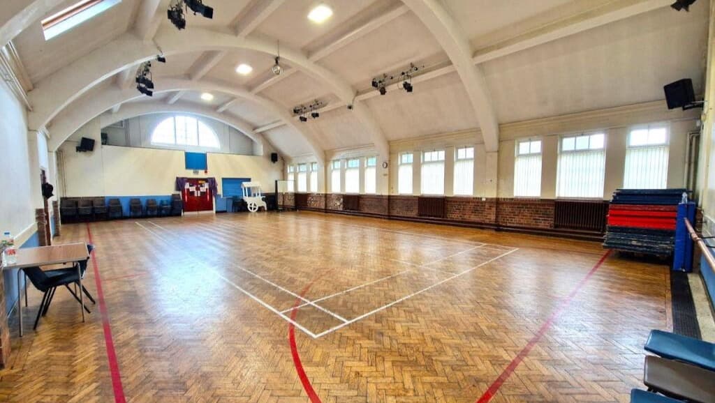 Spacious indoor gymnasium with wooden floors, high arched ceiling, stacked mats, and chairs along the walls. - Home Instead