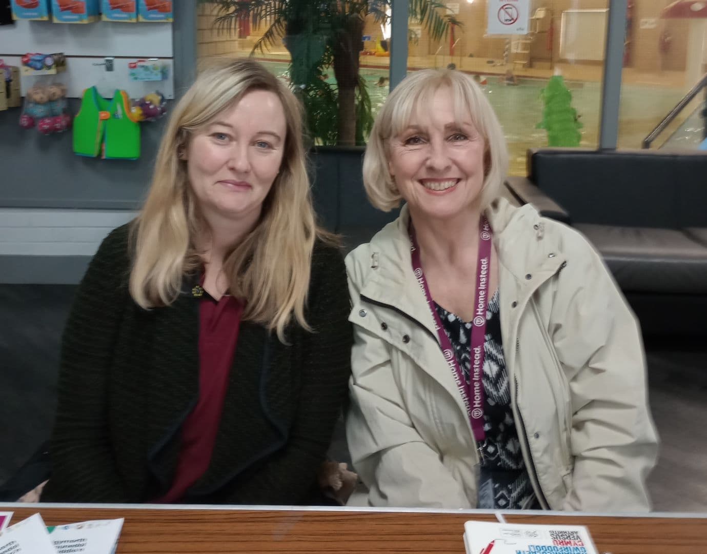 Two women smiling, seated at a table with leaflets, with a pool and play area in the background. - Home Instead