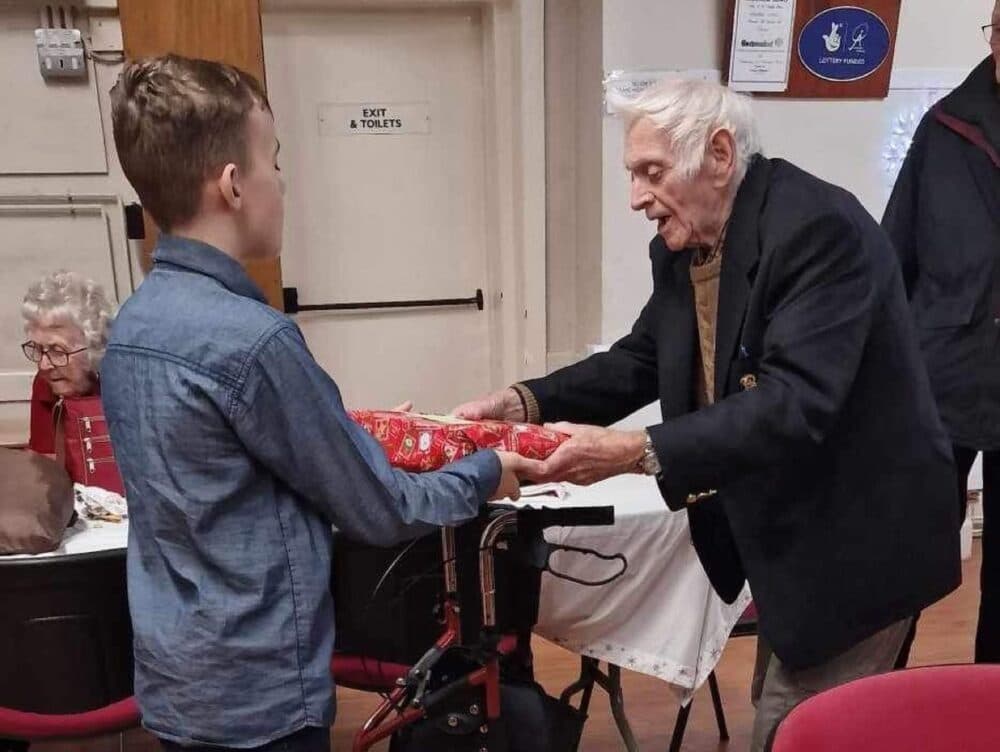 An elderly man hands a wrapped gift to a boy at a gathering, with others seated nearby. - Home Instead