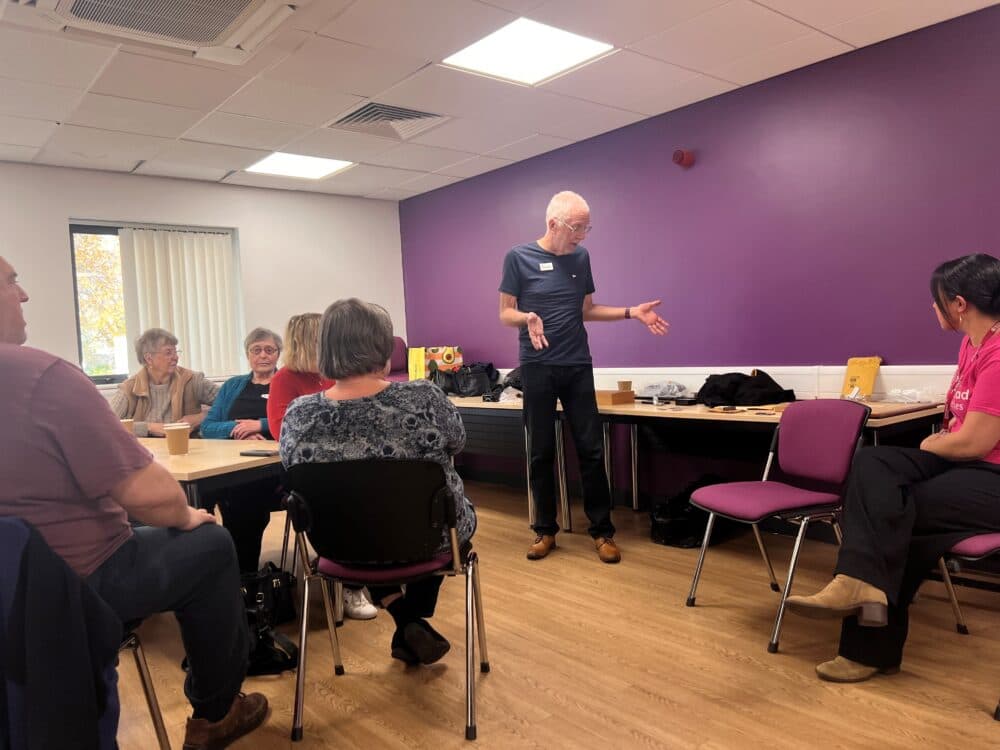 A man stands and speaks to a small group seated around tables in a room with purple walls. - Home Instead