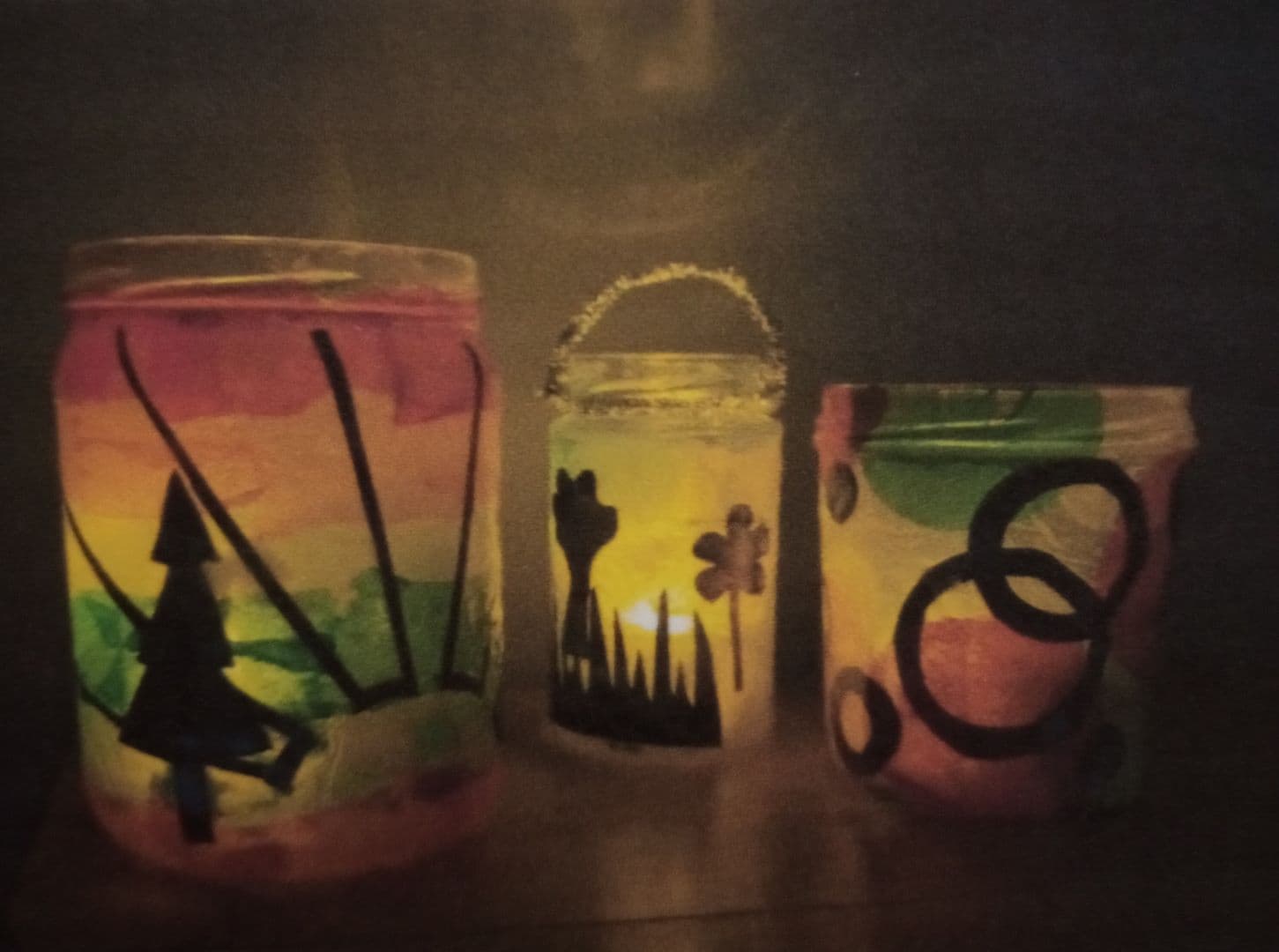 Three colorful, decorated glass jars with candles inside, glowing in the dark. - Home Instead