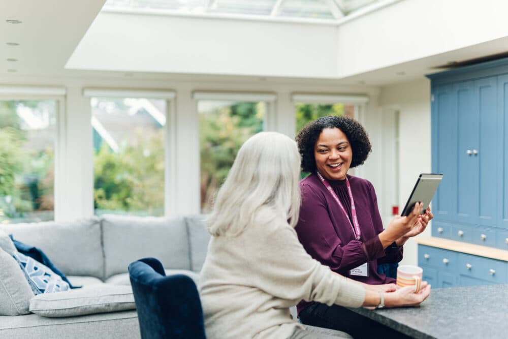 Two women sit together in a bright living room, smiling and looking at a tablet. - Home Instead
