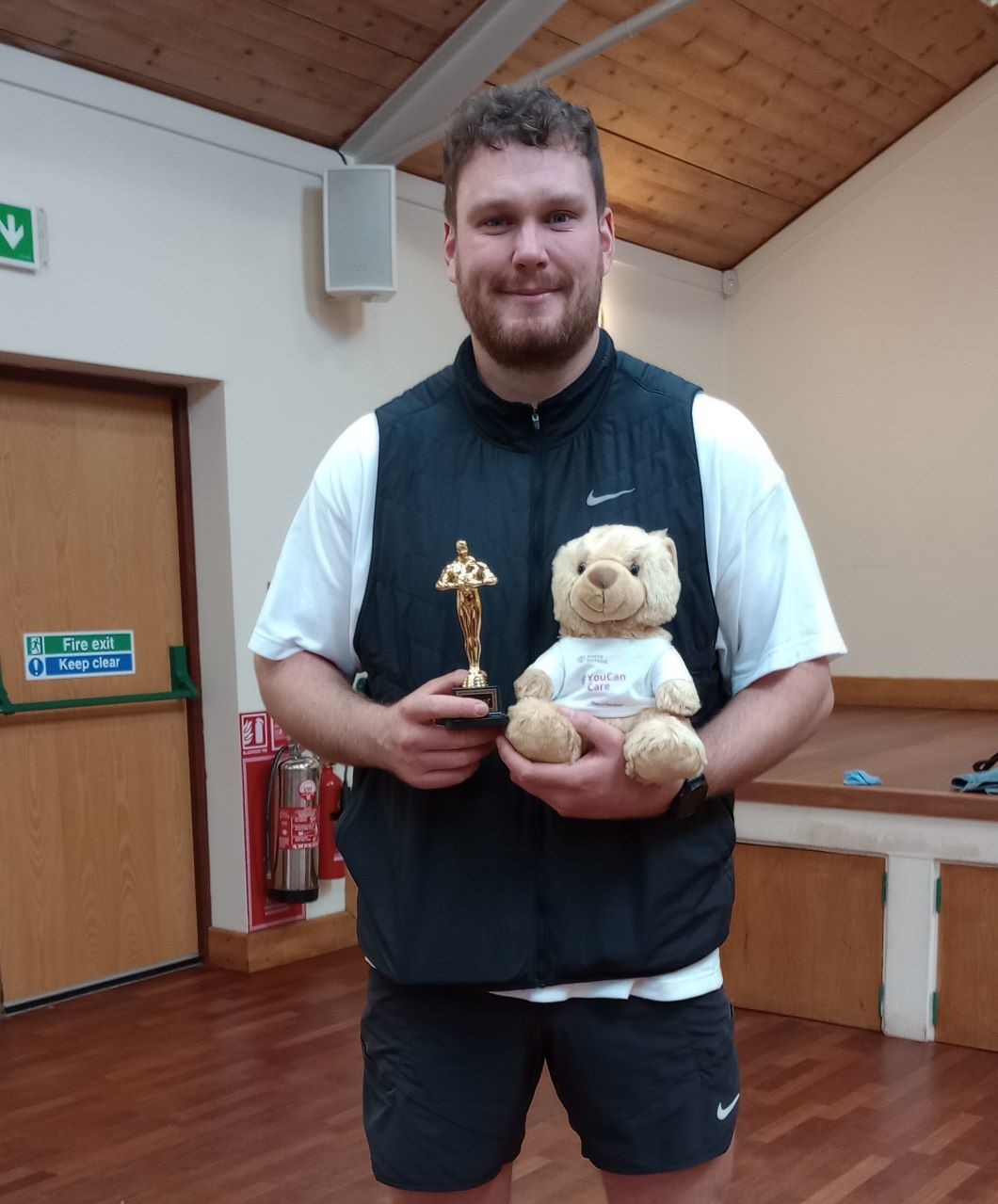 Man in sportswear holding a trophy and a teddy bear in a room with a wooden ceiling and fire exit sign. - Home Instead