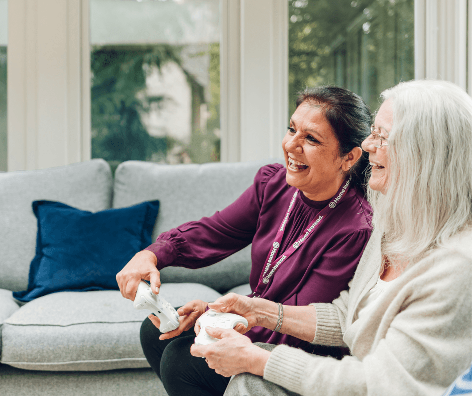 Two women smiling and playing video games together on a couch in a bright living room. - Home Instead