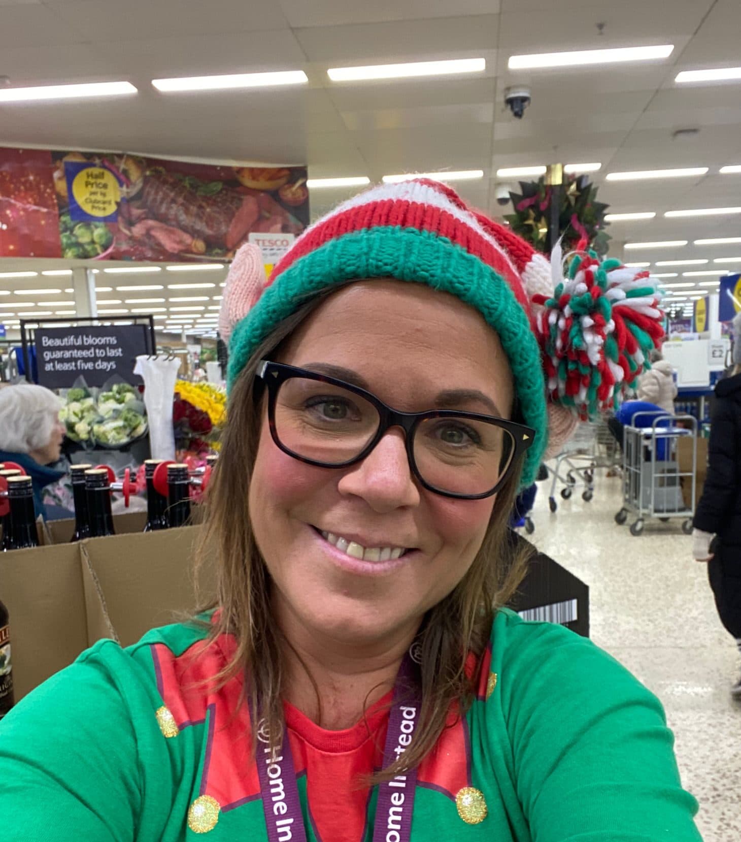 Smiling woman in glasses and elf hat takes a selfie inside a supermarket decorated for the holidays. - Home Instead