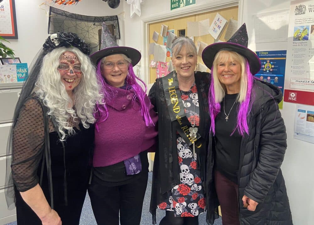 Four smiling women dressed in Halloween costumes, including witches' hats and wigs, pose together indoors. - Home Instead