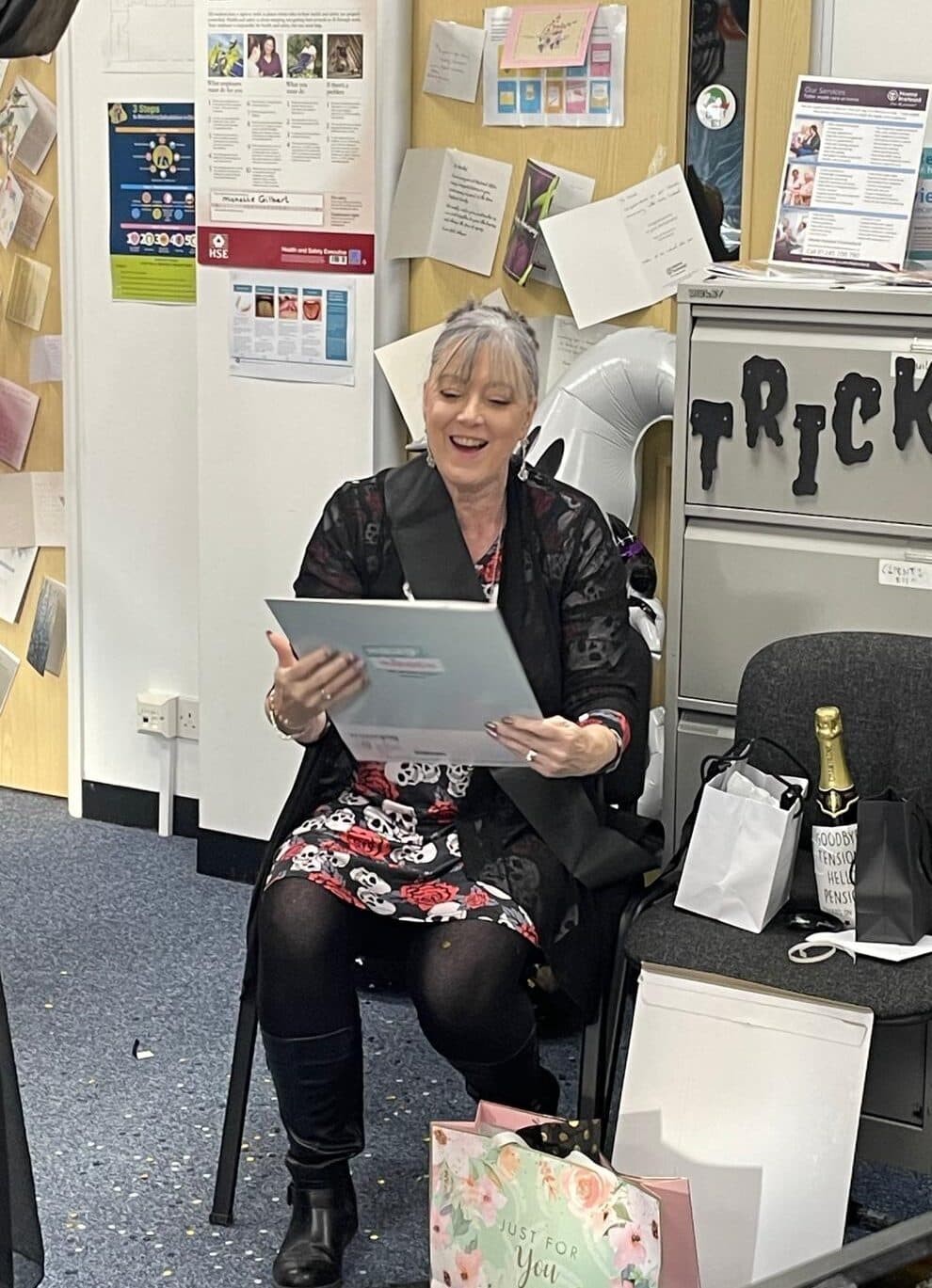 A smiling woman sits opening a large card, surrounded by gifts and decorations in an office setting. - Home Instead