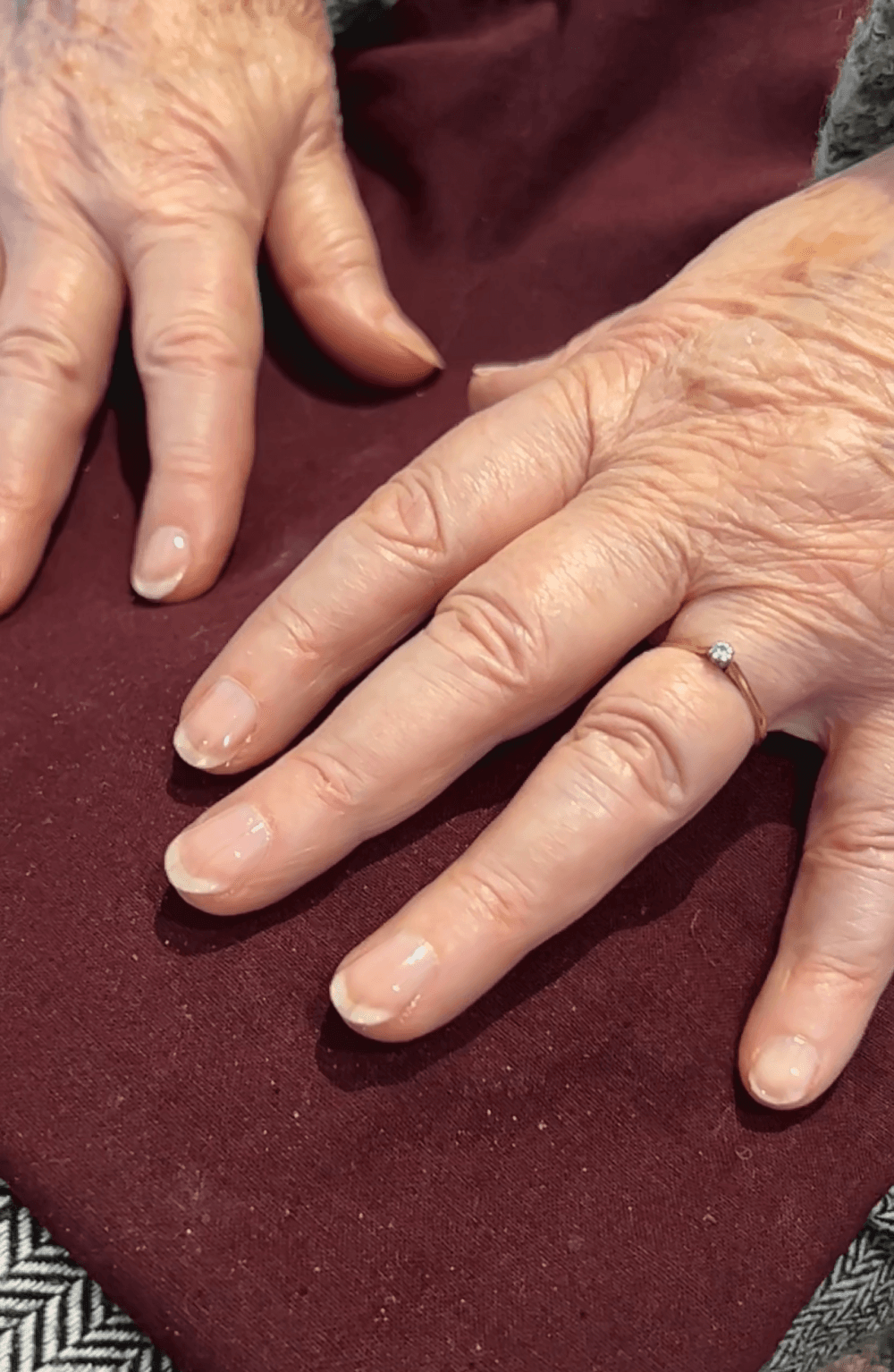 Elderly hands with natural nails rest on a maroon surface; one hand has a gold ring with a small stone. - Home Instead
