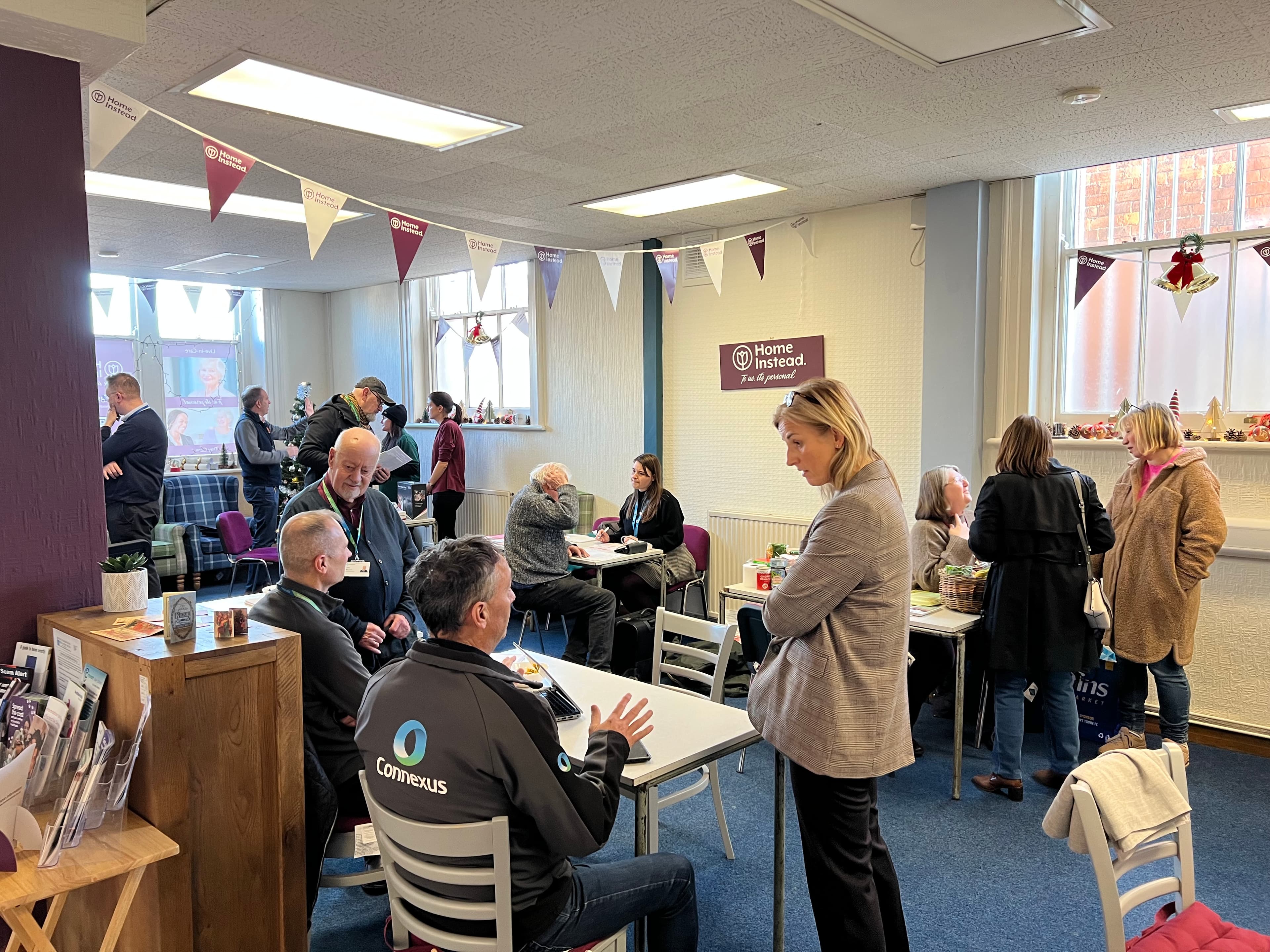 People chatting and drinking at a bright, cozy community café decorated with bunting and autumn ornaments. - Home Instead
