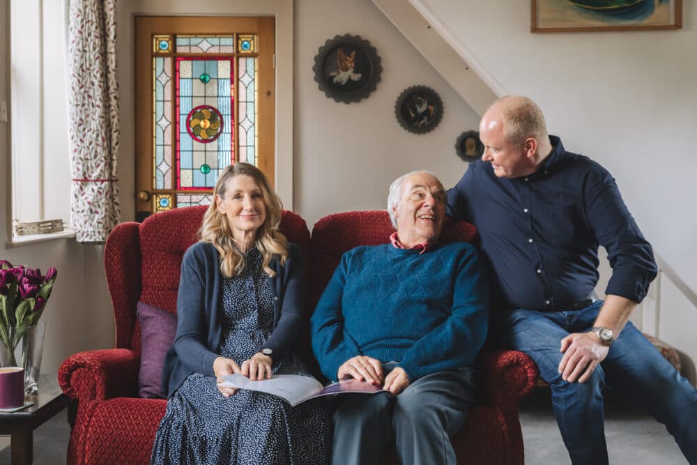 Three people sit together on a red sofa, smiling and talking in a cozy, warmly lit living room. - Home Instead