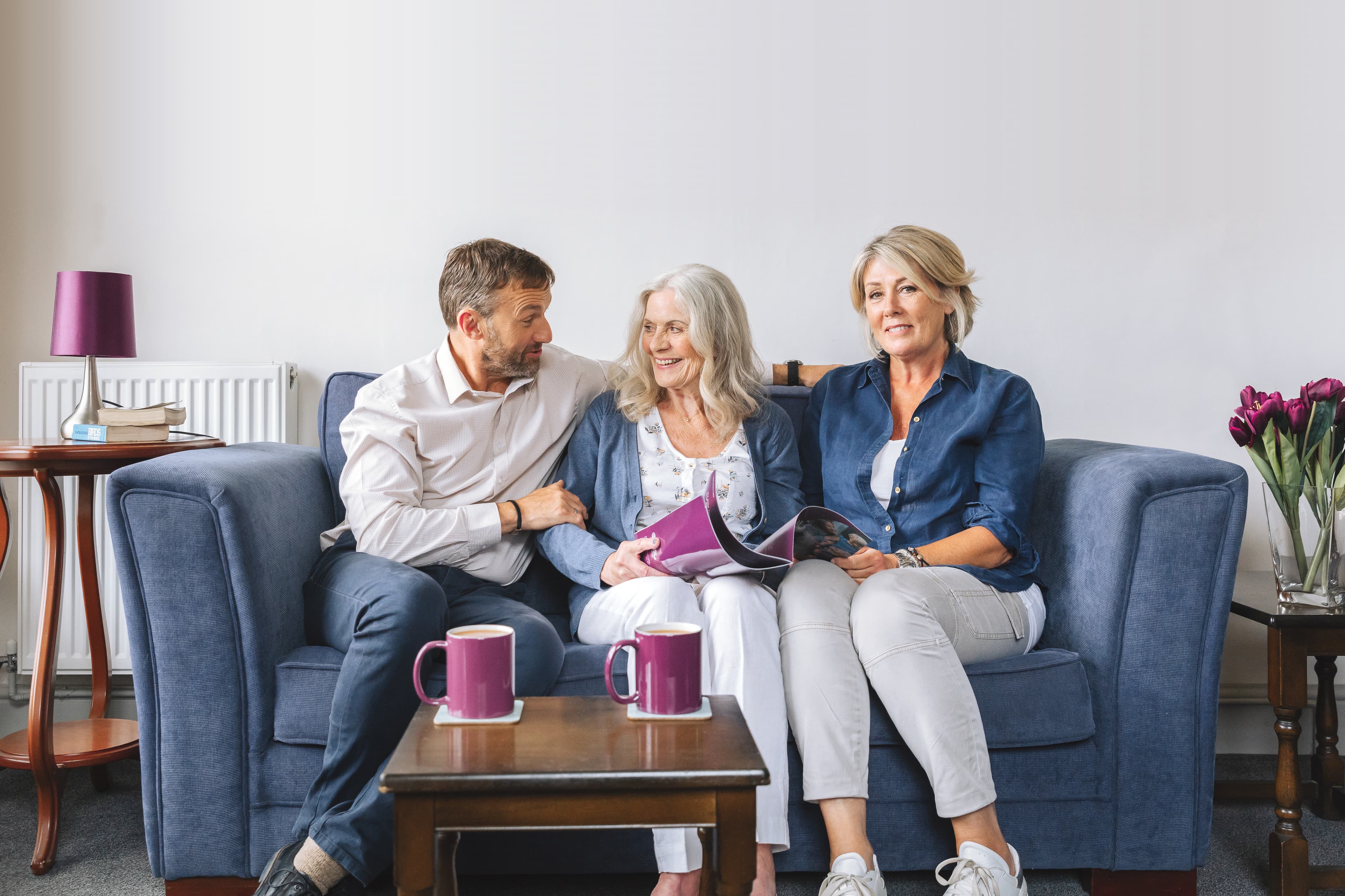 Three smiling adults sit together on a blue sofa with coffee mugs on a table in front of them. - Home Instead