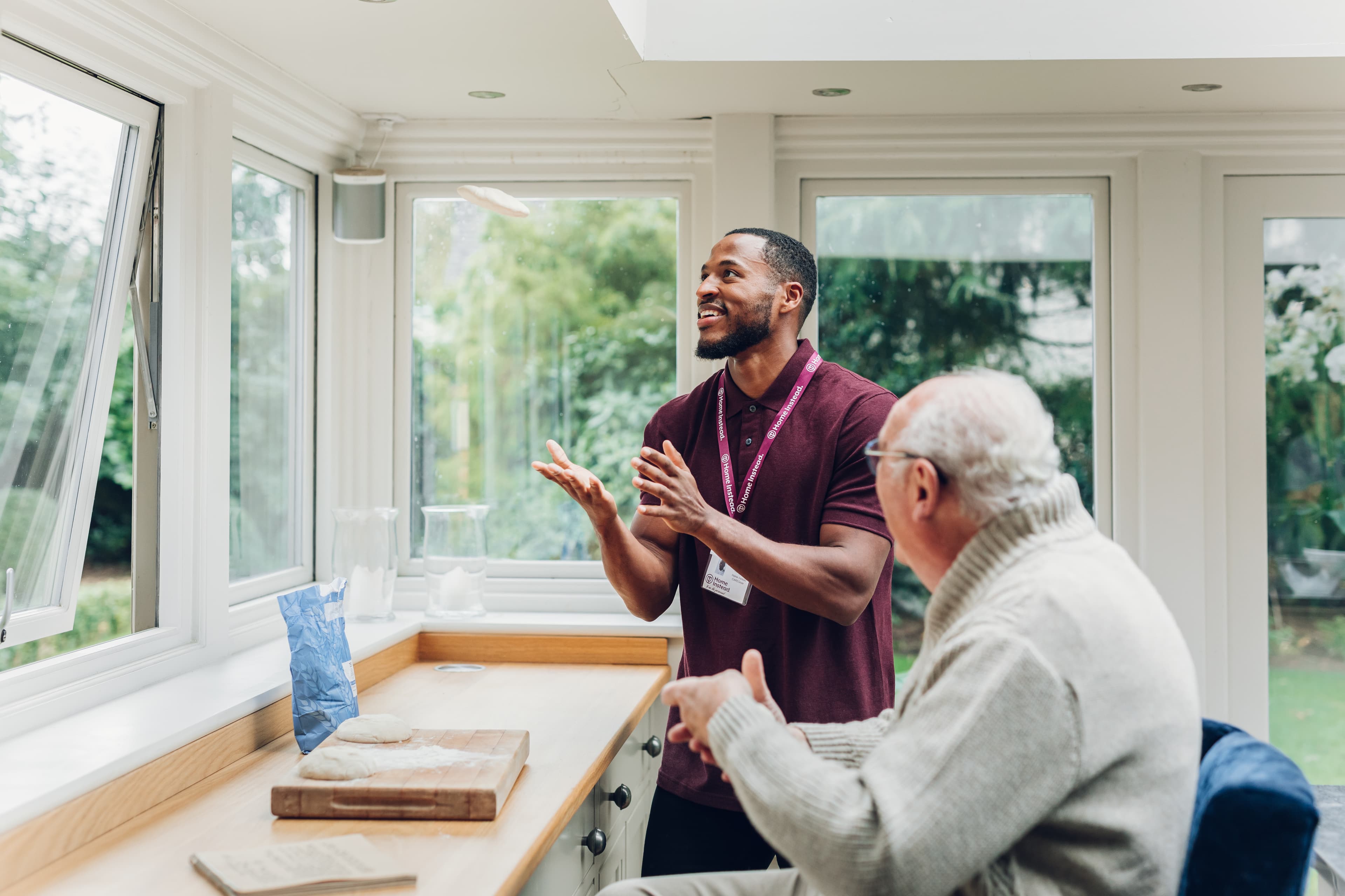 A young man and an older man talk and smile together in a bright kitchen with large windows. - Home Instead
