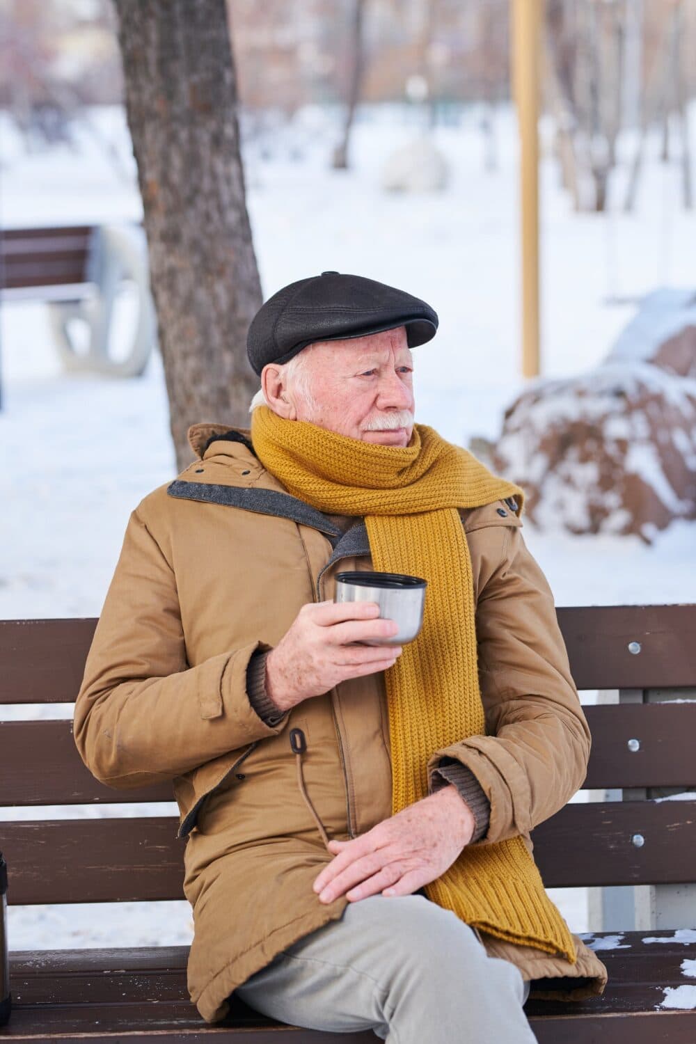 Elderly man in winter clothes sits on a bench outdoors, holding a mug, with snow in the background. - Home Instead