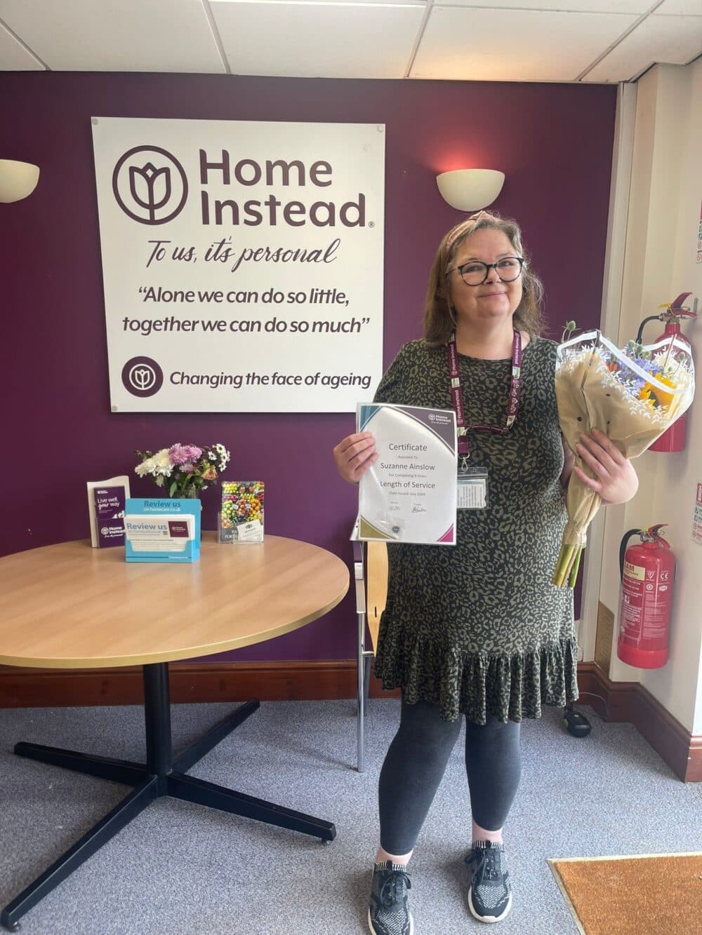 Smiling woman holding flowers and a certificate stands in front of a "Home Instead" sign in an office. - Home Instead