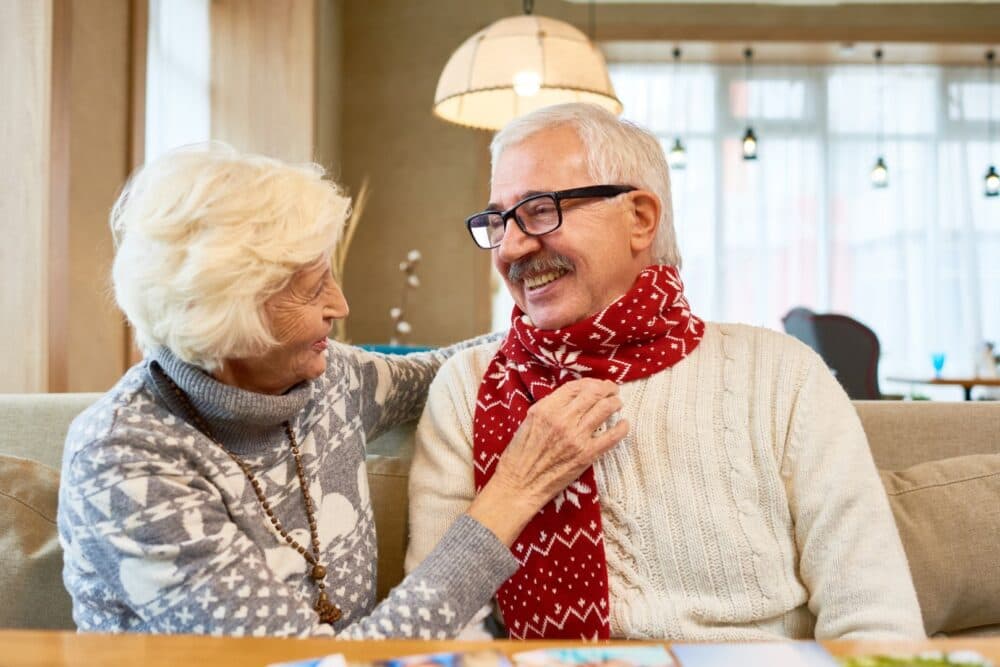 Elderly woman smiling as she adjusts a red scarf on an elderly man sitting beside her indoors. - Home Instead