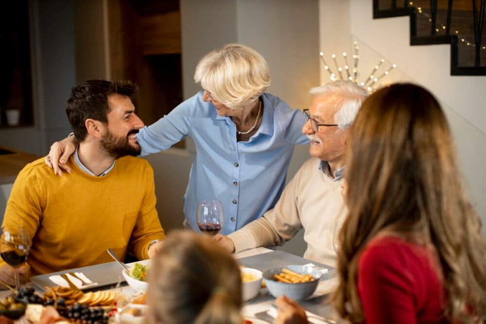 Four people enjoying a meal together, smiling and talking around a dining table in a cozy home setting. - Home Instead