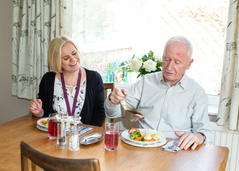 An elderly man and a woman sit at a table, sharing a meal and smiling in a bright dining room. - Home Instead