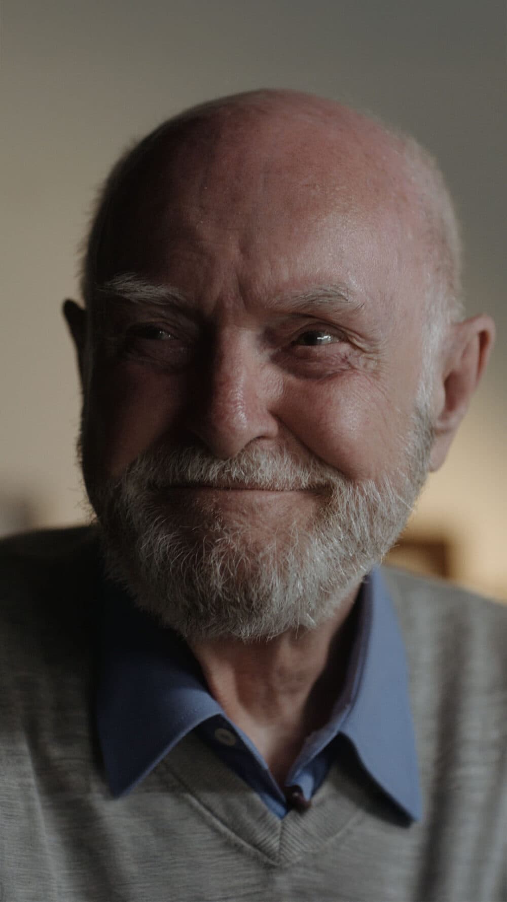 An elderly man with a beard smiles warmly, wearing a blue shirt and light sweater. - Home Instead