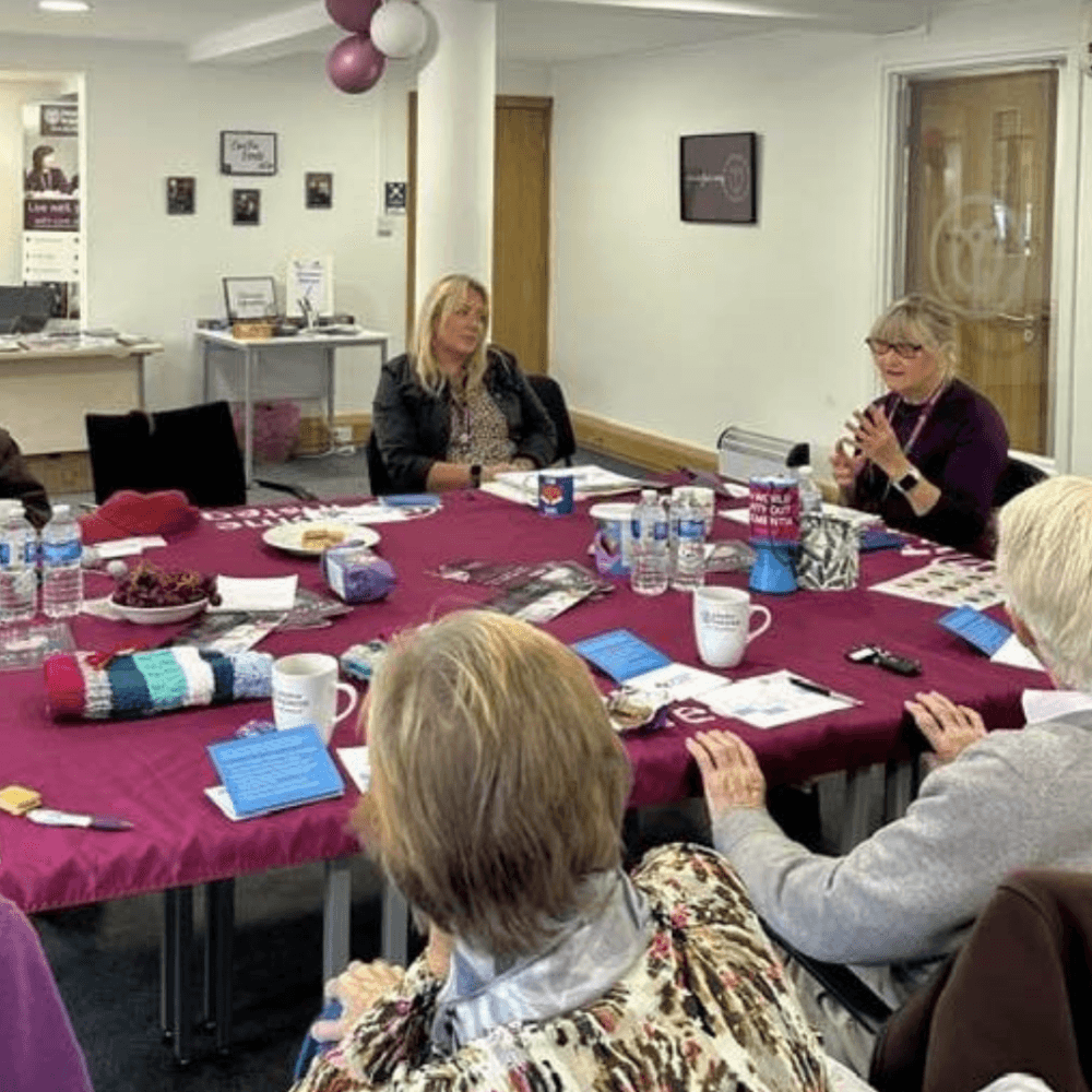 Six women sit around a table with papers, drinks, and snacks, having a discussion in a meeting room. - Home Instead