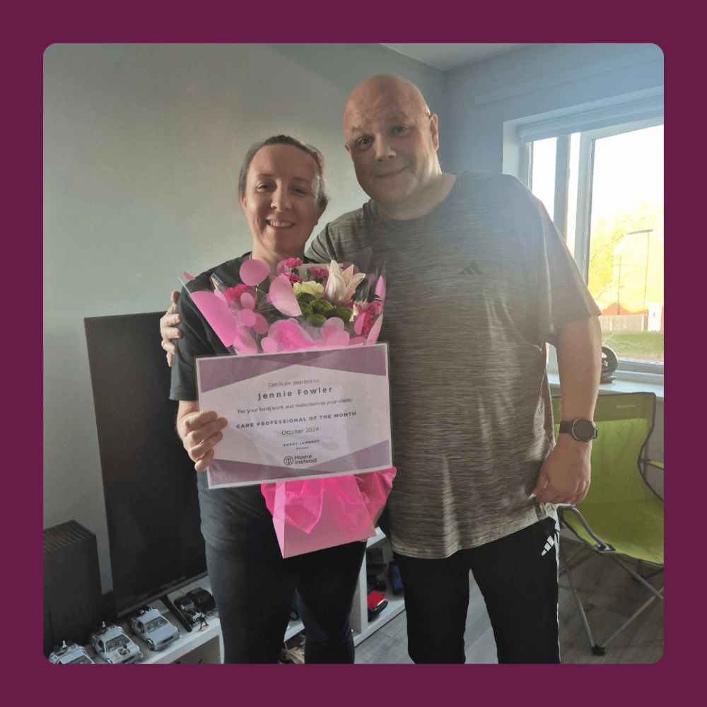 Two people smiling; one holds a bouquet of flowers and a certificate that reads "Jenale Fowler. - Home Instead