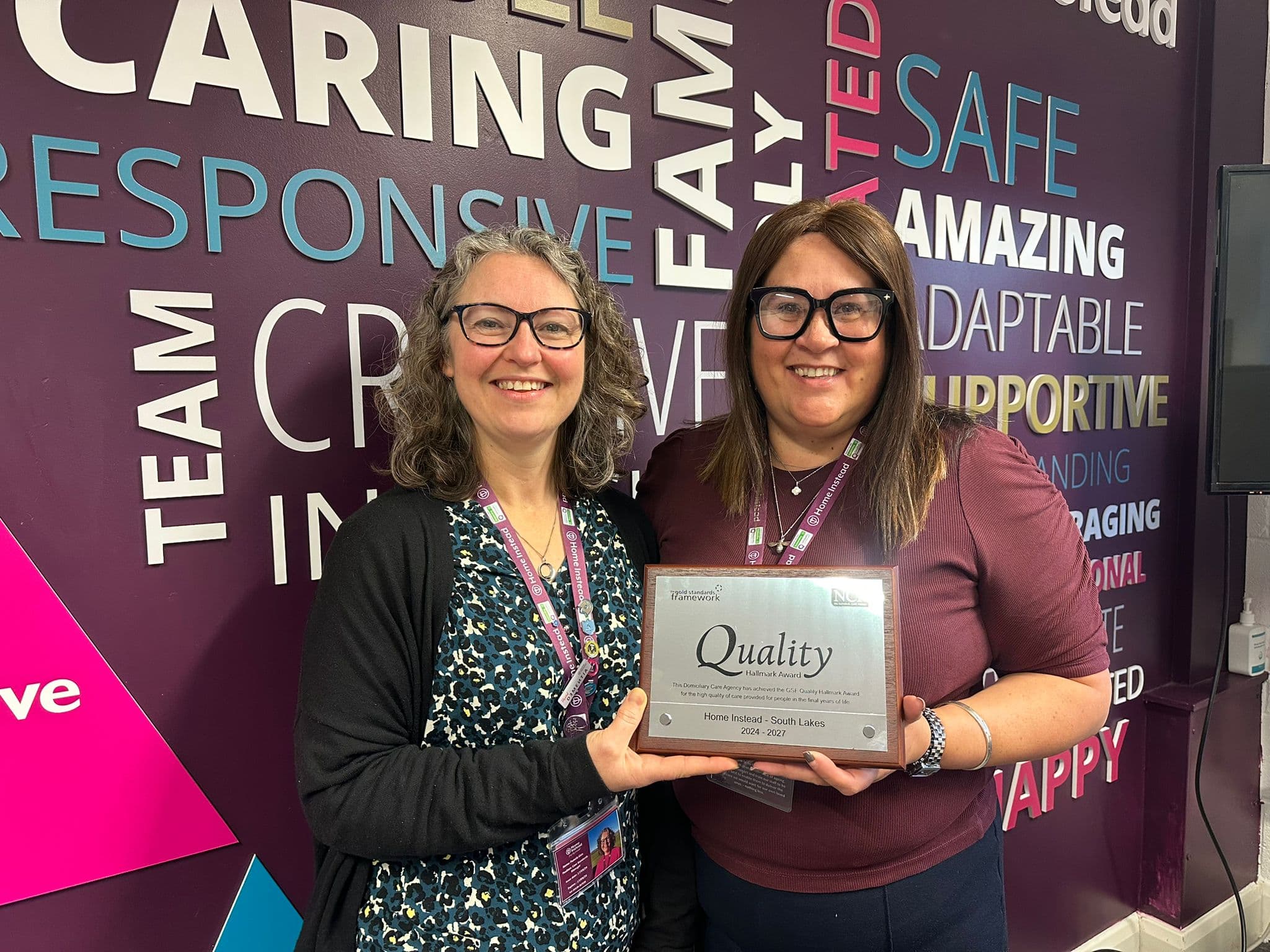 Two women smiling and holding a "Quality" award plaque in front of a colorful, word-filled wall. - Home Instead