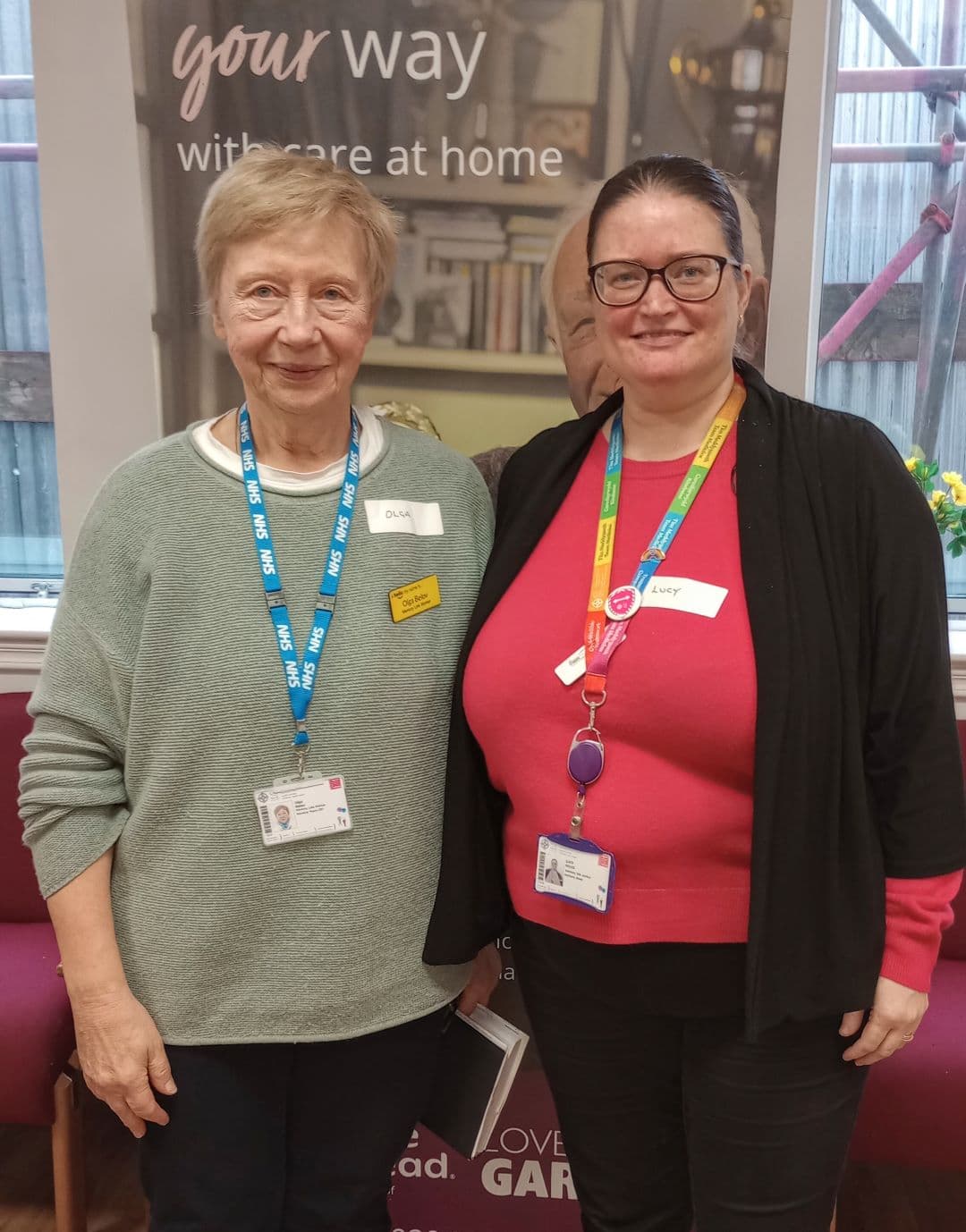 Two women with name badges and lanyards stand side by side, smiling indoors at a care facility. - Home Instead