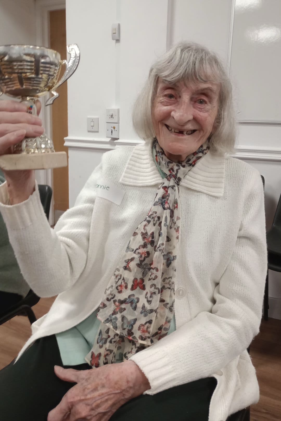 Elderly woman smiling and holding up a trophy, wearing a white sweater and floral scarf. - Home Instead