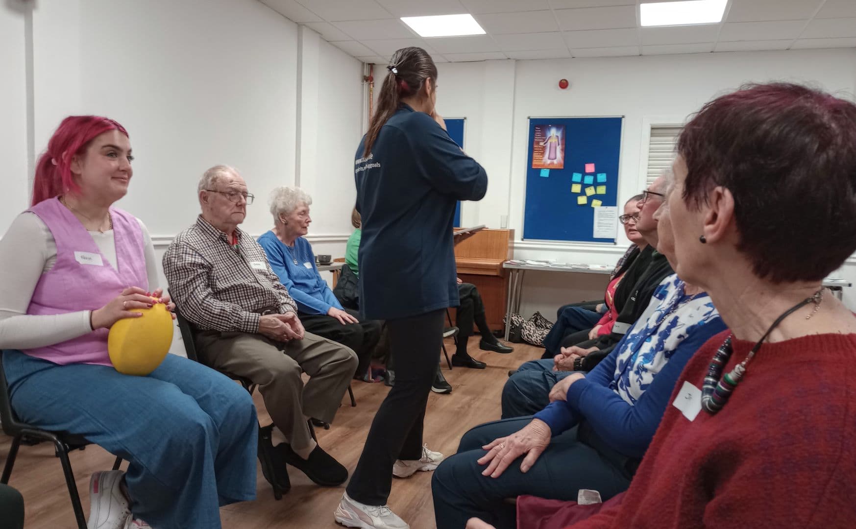A group of people sit in a circle indoors, talking; one woman holds a yellow balloon. - Home Instead