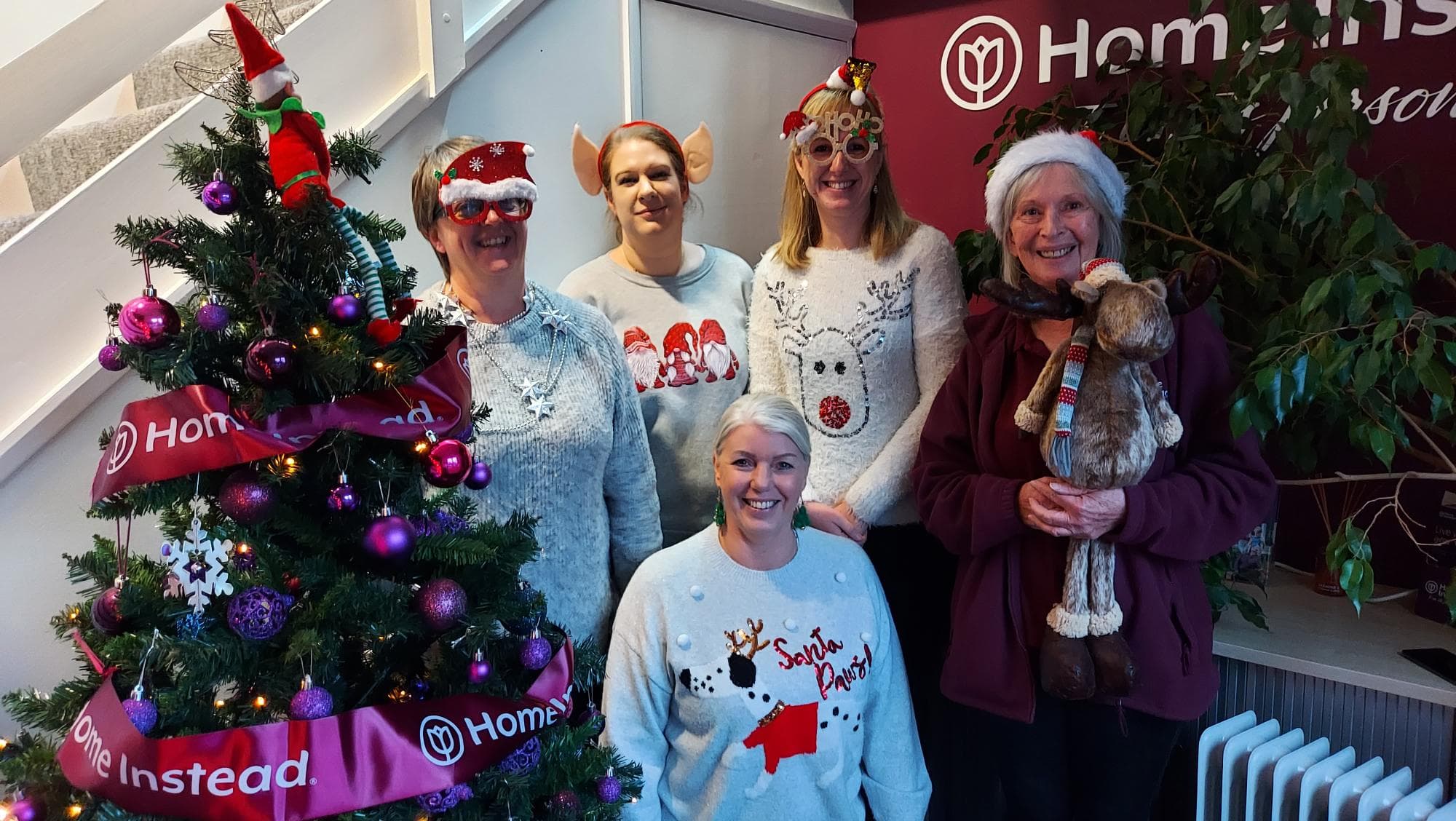 Five women in festive sweaters and hats pose by a decorated Christmas tree in an office setting. - Home Instead