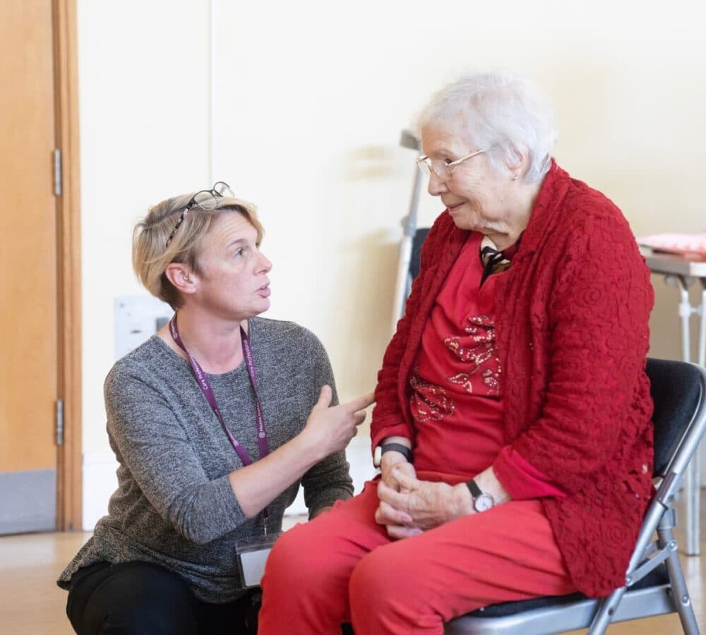 A carer with eyeglasses on his head kneeling while speaking to a senior woman with grey hair, sitting down and wearing read