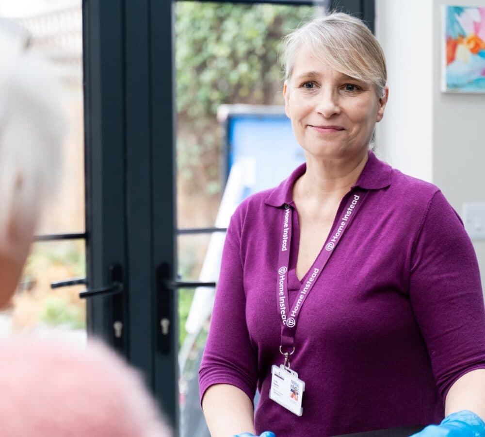 A lady carer wearing an ID and gloves smiling while at the kitchen with glass doors and while talking to someone