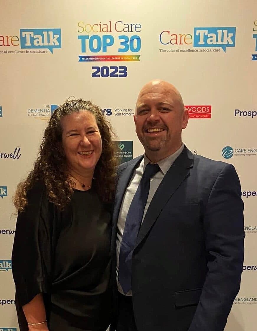A smiling man in a suit and woman in a black dress pose at a Social Care Top 30 event backdrop. - Home Instead