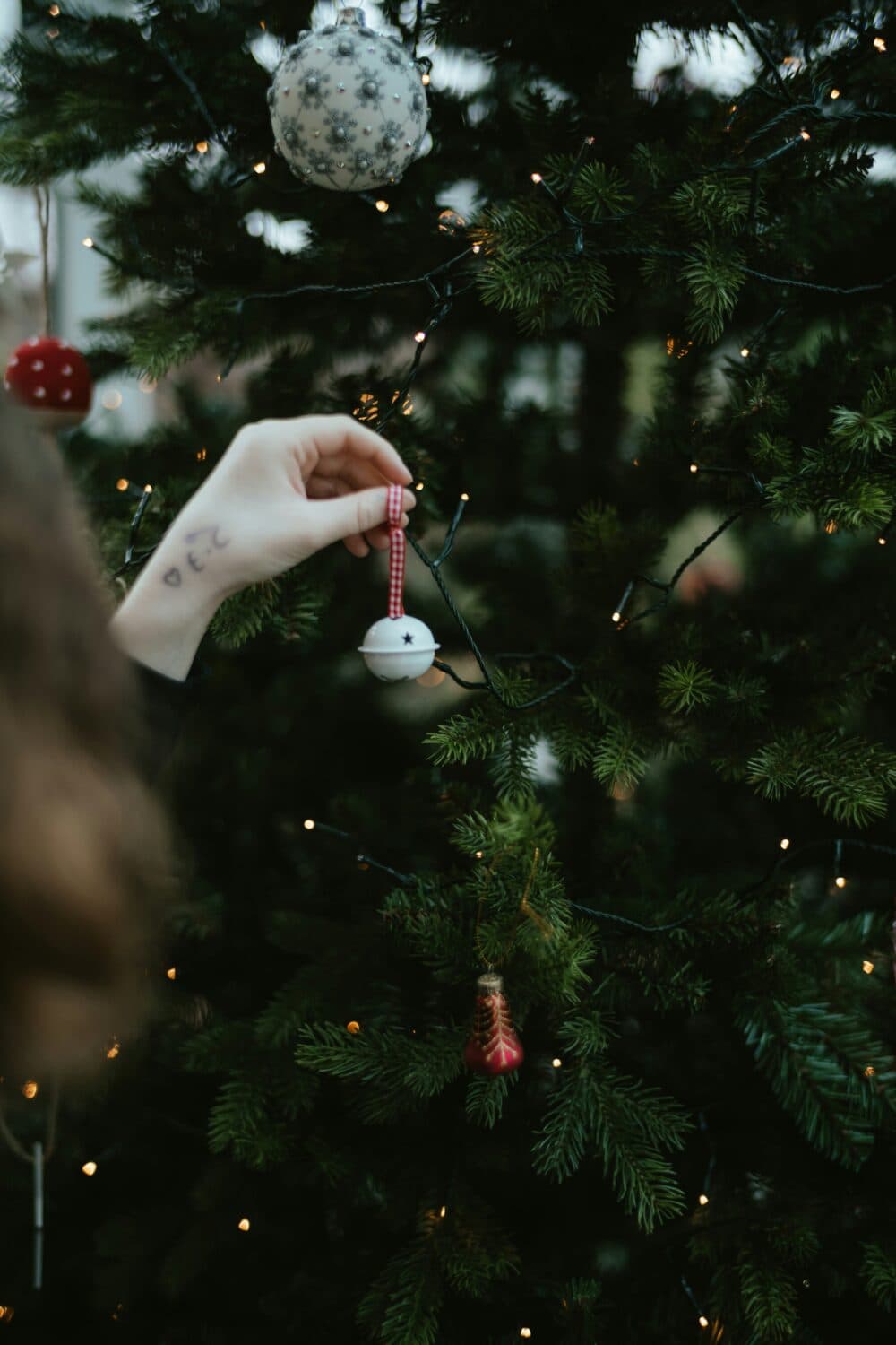 A hand hangs a white bauble on a Christmas tree decorated with lights and ornaments. - Home Instead