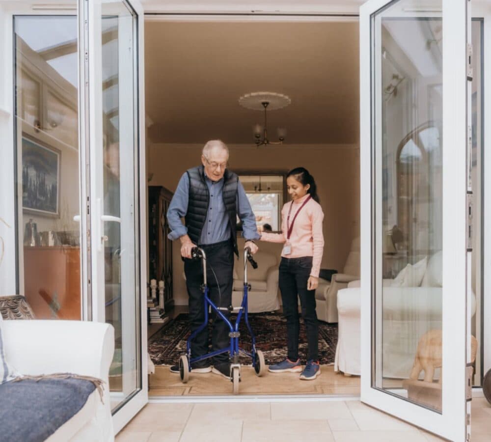 A carer assisting a senior man using a walker while walking out the door of the ouse