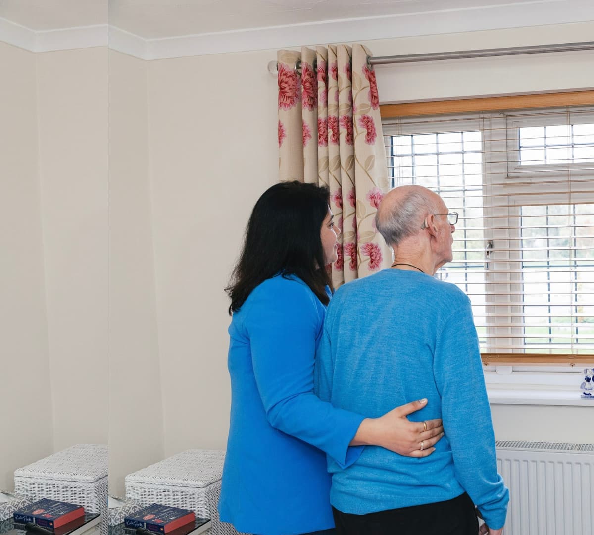 Carer helping a Senior man both wearing blue looking at the window inside a room