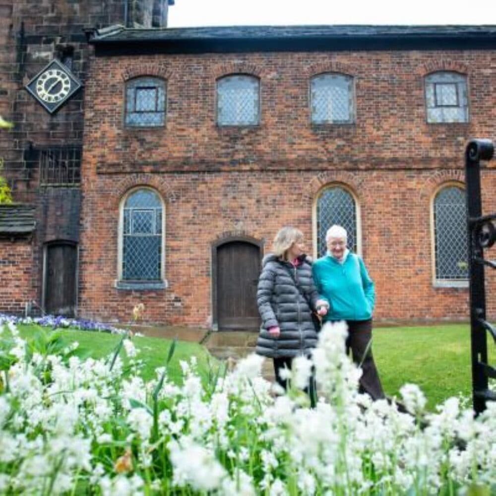 Two women walk and smile outside a brick building with a clock, behind a bed of white flowers. - Home Instead