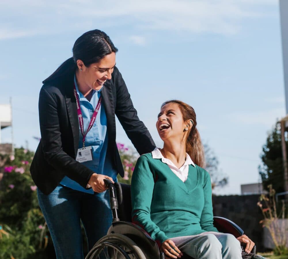 A caregiver smiles while pushing a young woman in a wheelchair outdoors on a sunny day. - Home Instead