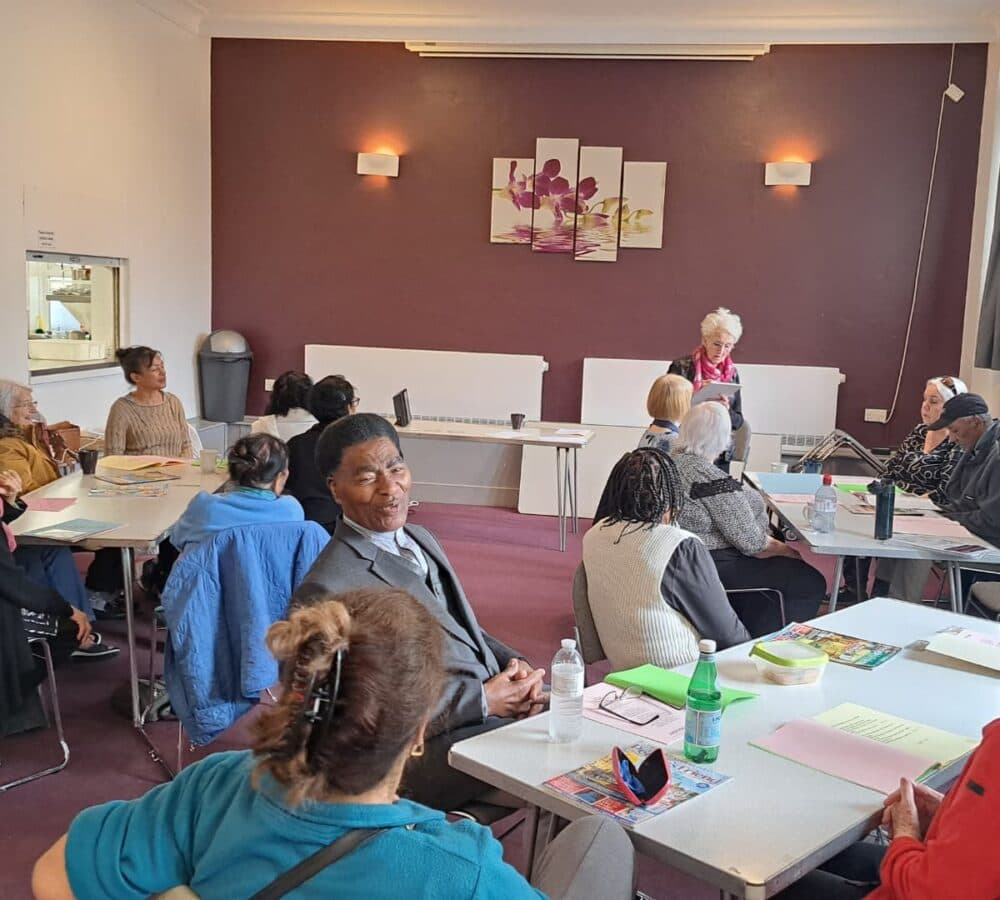 Group of elderly sitting down inside a room with tables and there are papers and bottles on the table, everyone is chatting
