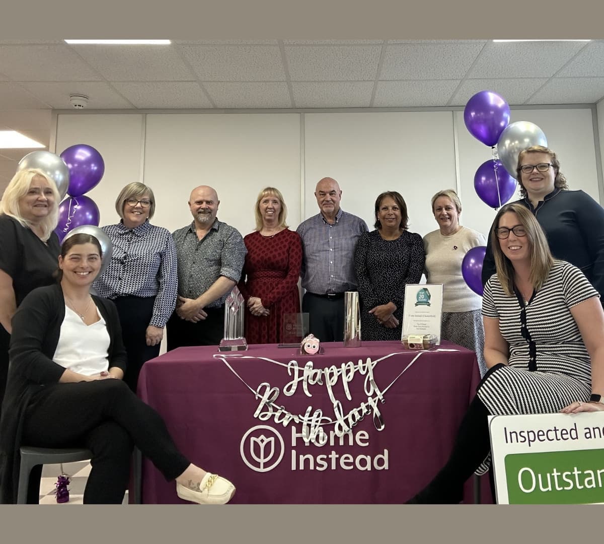 Group of men and women all happy and smiling with purple balloons and a Home Instead and Happy Birthday banner