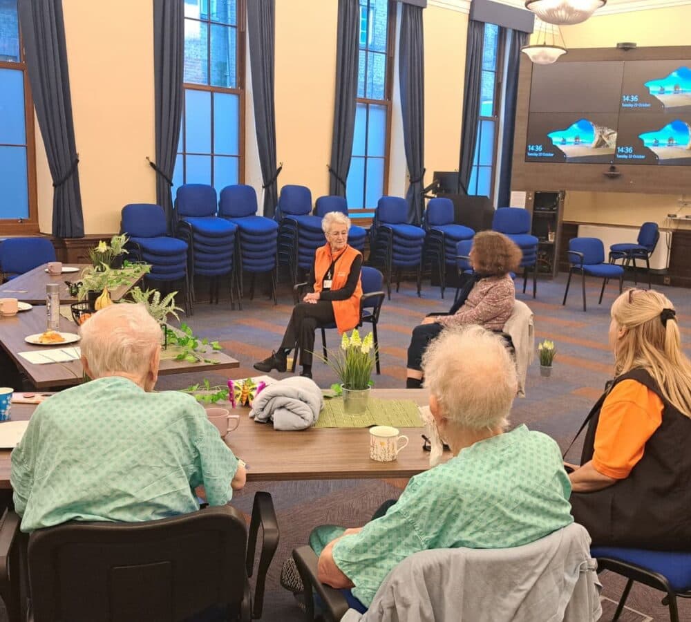 Group of seniors chatting inside a big room while sitting down two of them are wearing green and two of them are wearing orange with several chairs in the background